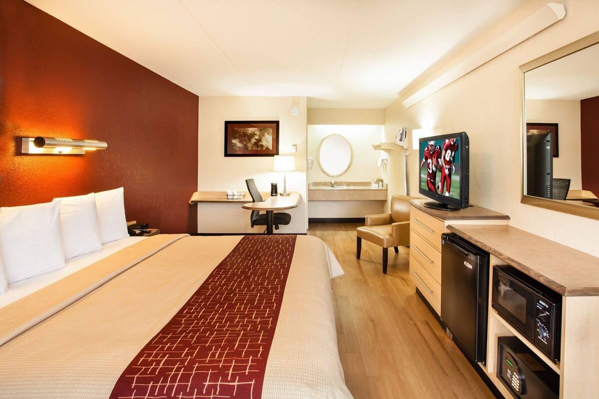 A hotel room with a bed, a desk with a chair, a television, a mirror, and a bathroom.