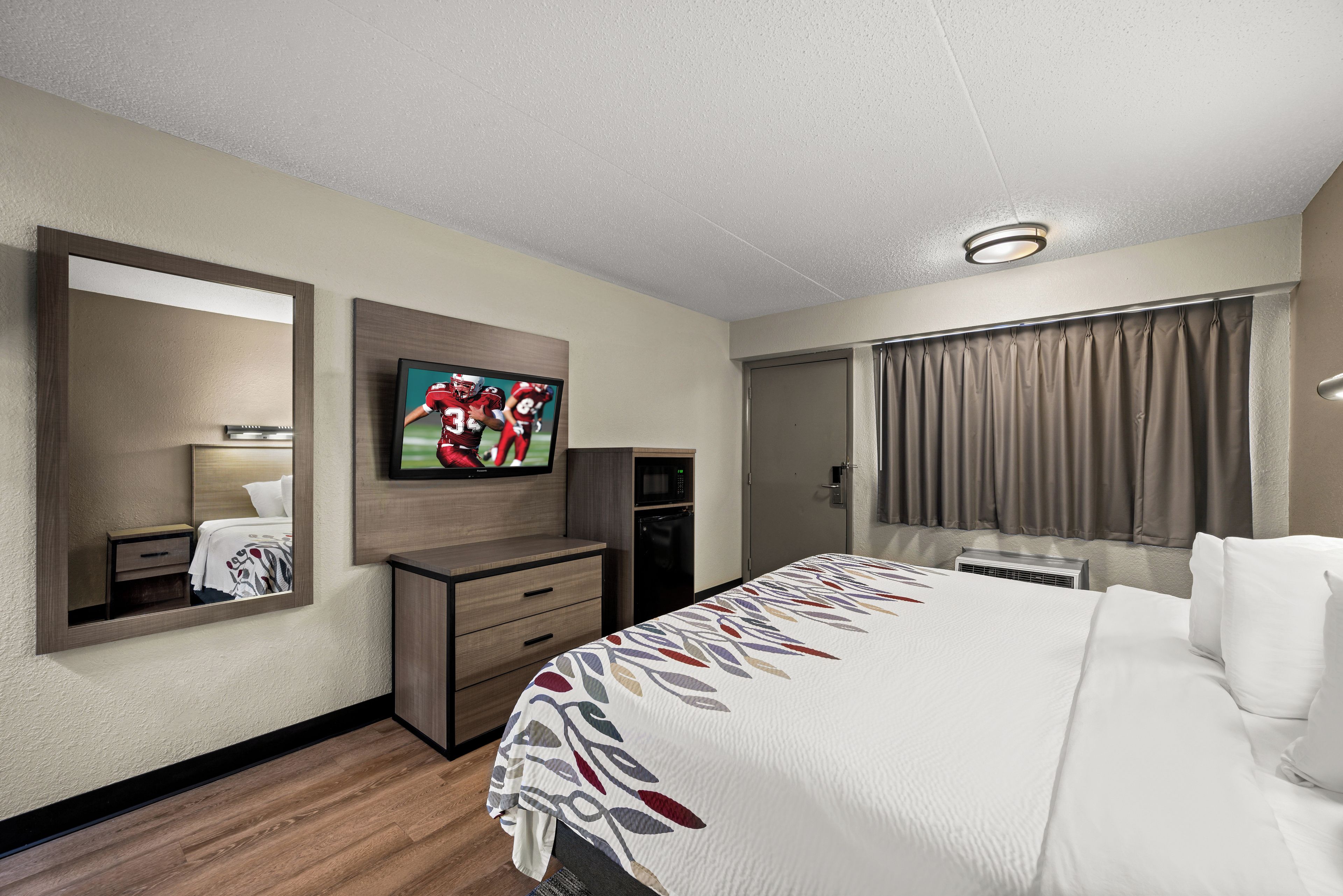 Una habitación de hotel con cama, televisión, escritorio, espejo y ventana con cortinas.