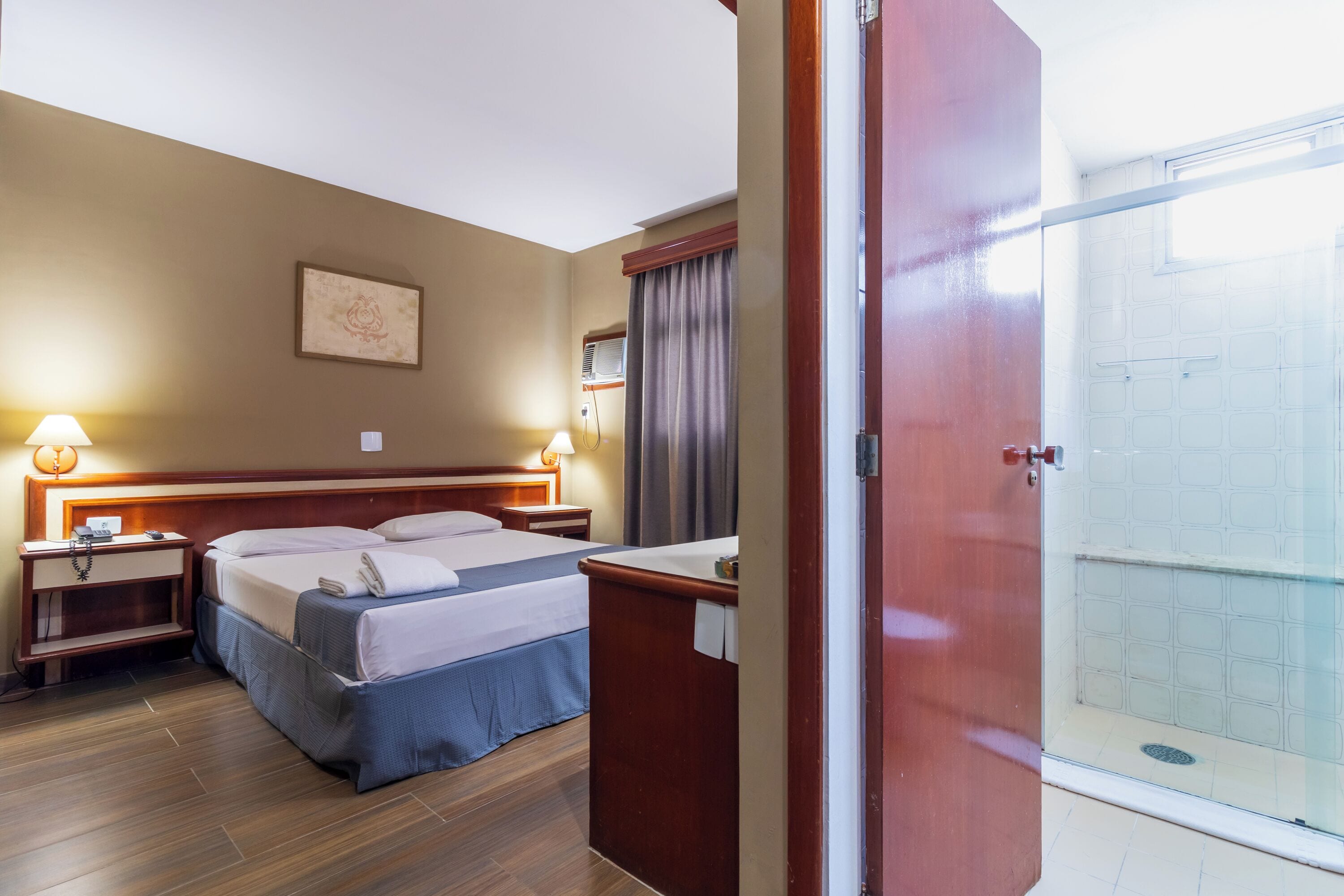 Habitación de hotel con cama, mesitas de noche, escritorio, zona de ducha y vistas al exterior.