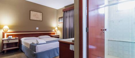 Habitación de hotel con cama, mesitas de noche, escritorio, zona de ducha y vistas al exterior.