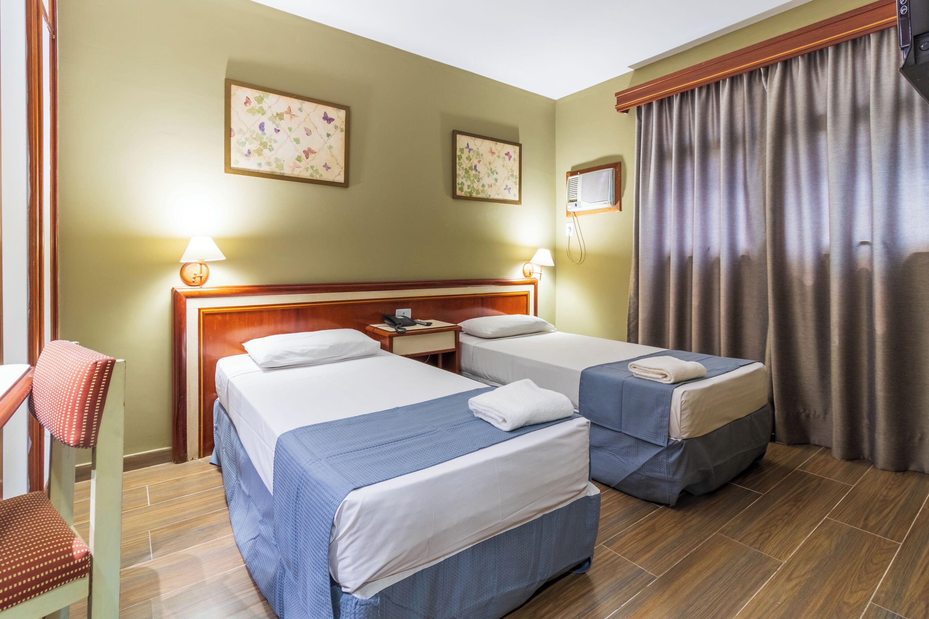 A hotel room with two beds, a wooden headboard, a desk with a lamp, and a window with curtains.