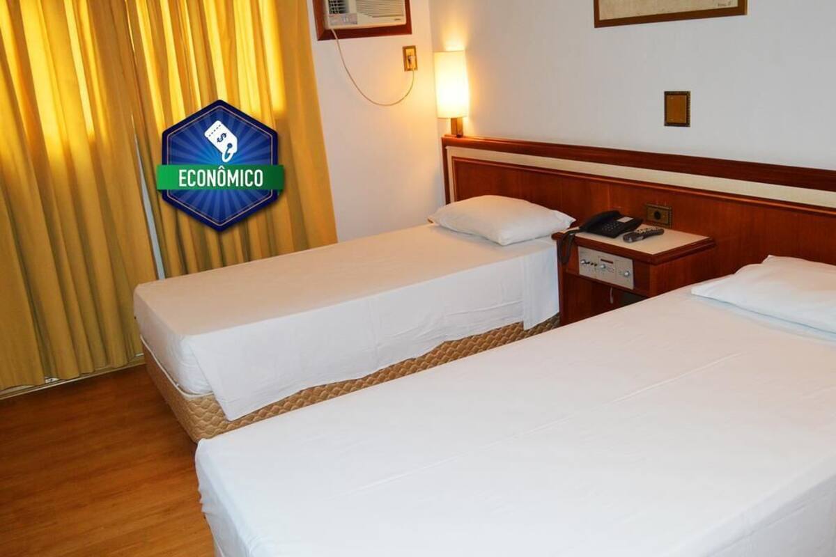 Quarto de hotel com duas camas, uma mesa de cabeceira com telefone e uma luminária na parede.