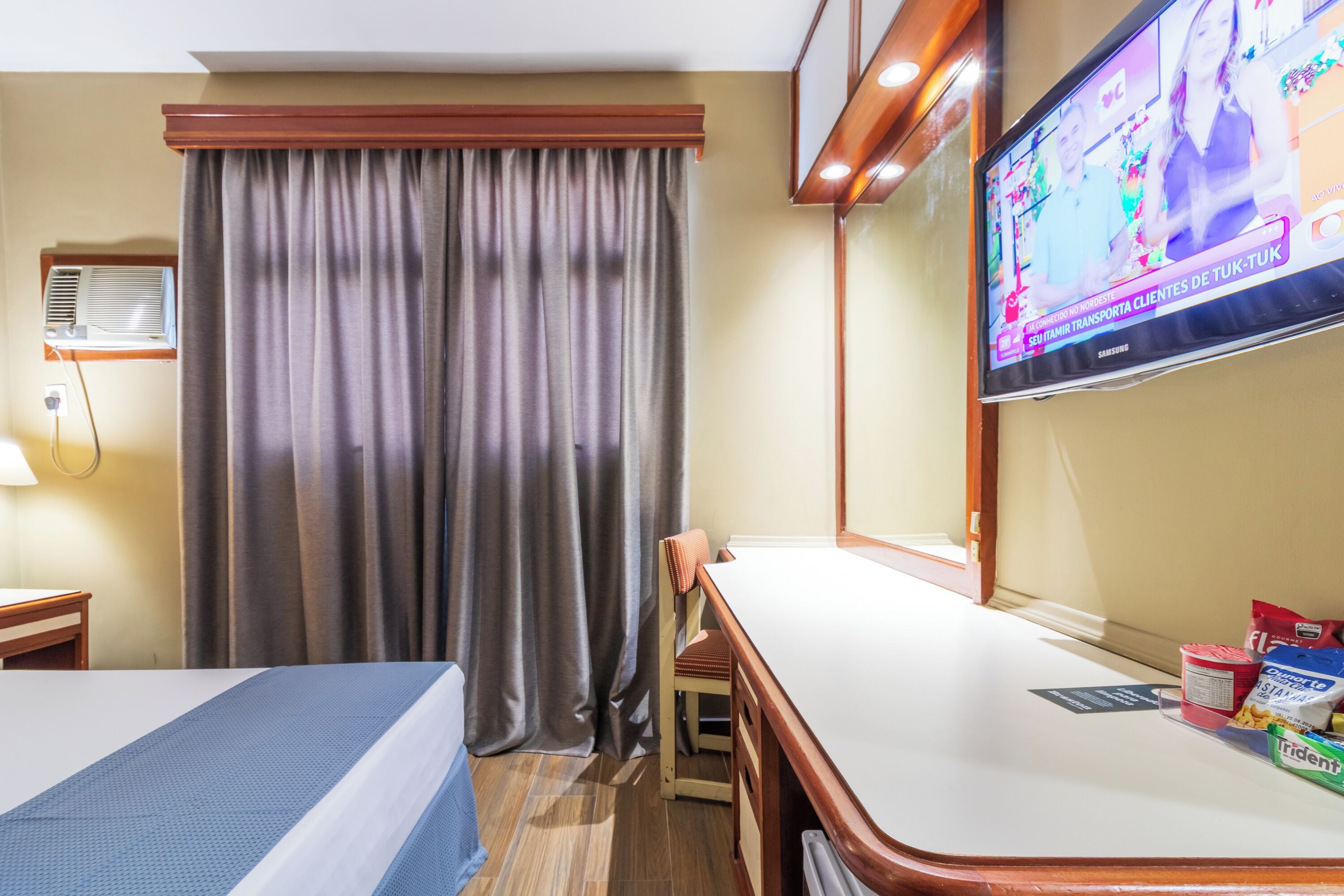 Una habitación de hotel con cama, escritorio, silla, televisor y ventana con cortinas.