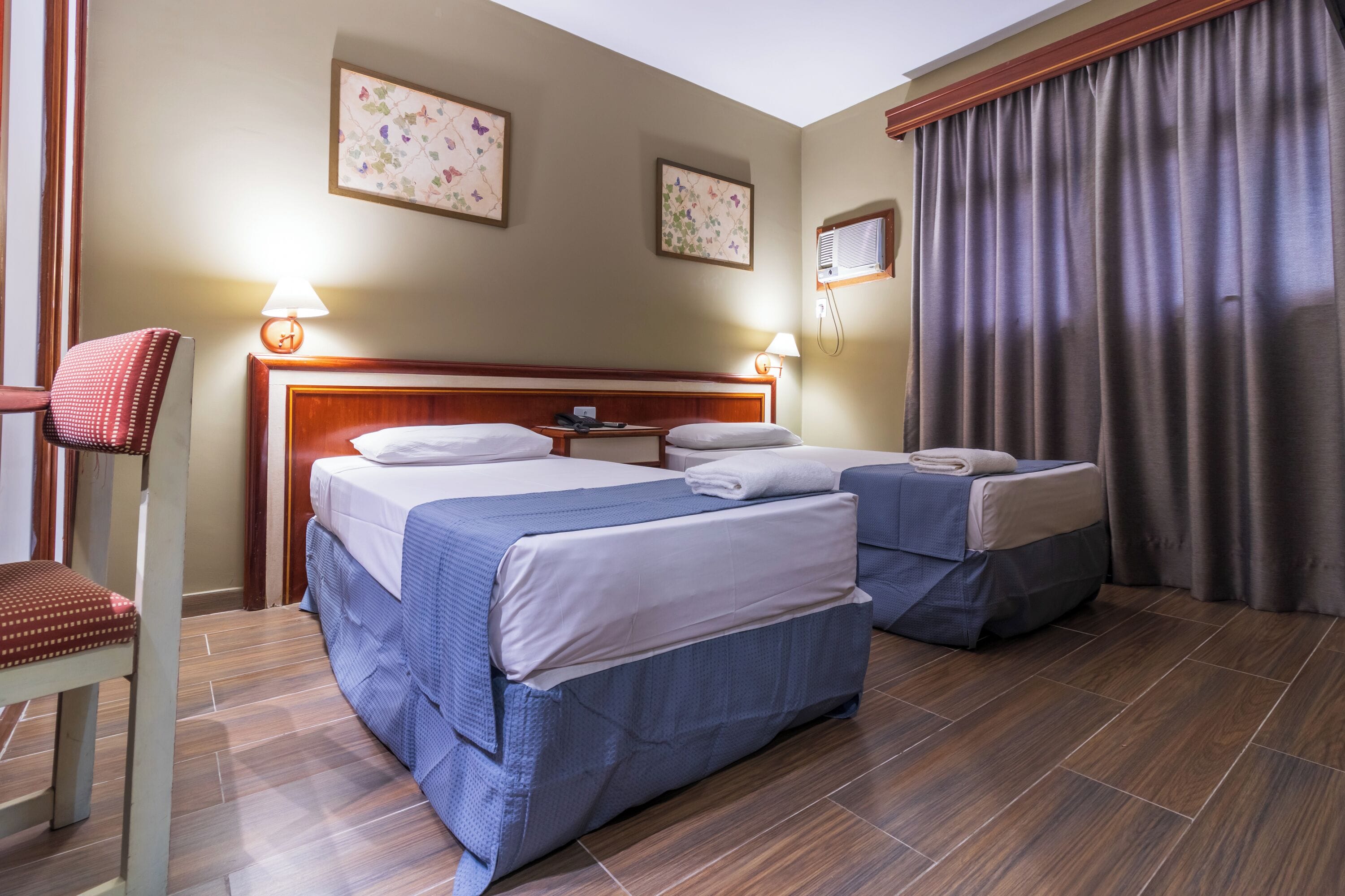 Quarto de hotel com duas camas, uma cadeira, uma mesa de cabeceira e uma janela com cortinas.