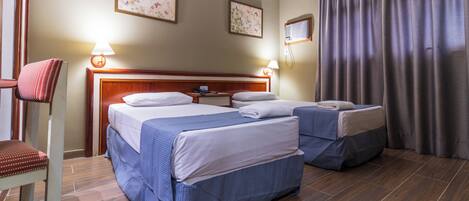 Quarto de hotel com duas camas, uma cadeira, uma mesa de cabeceira e uma janela com cortinas.