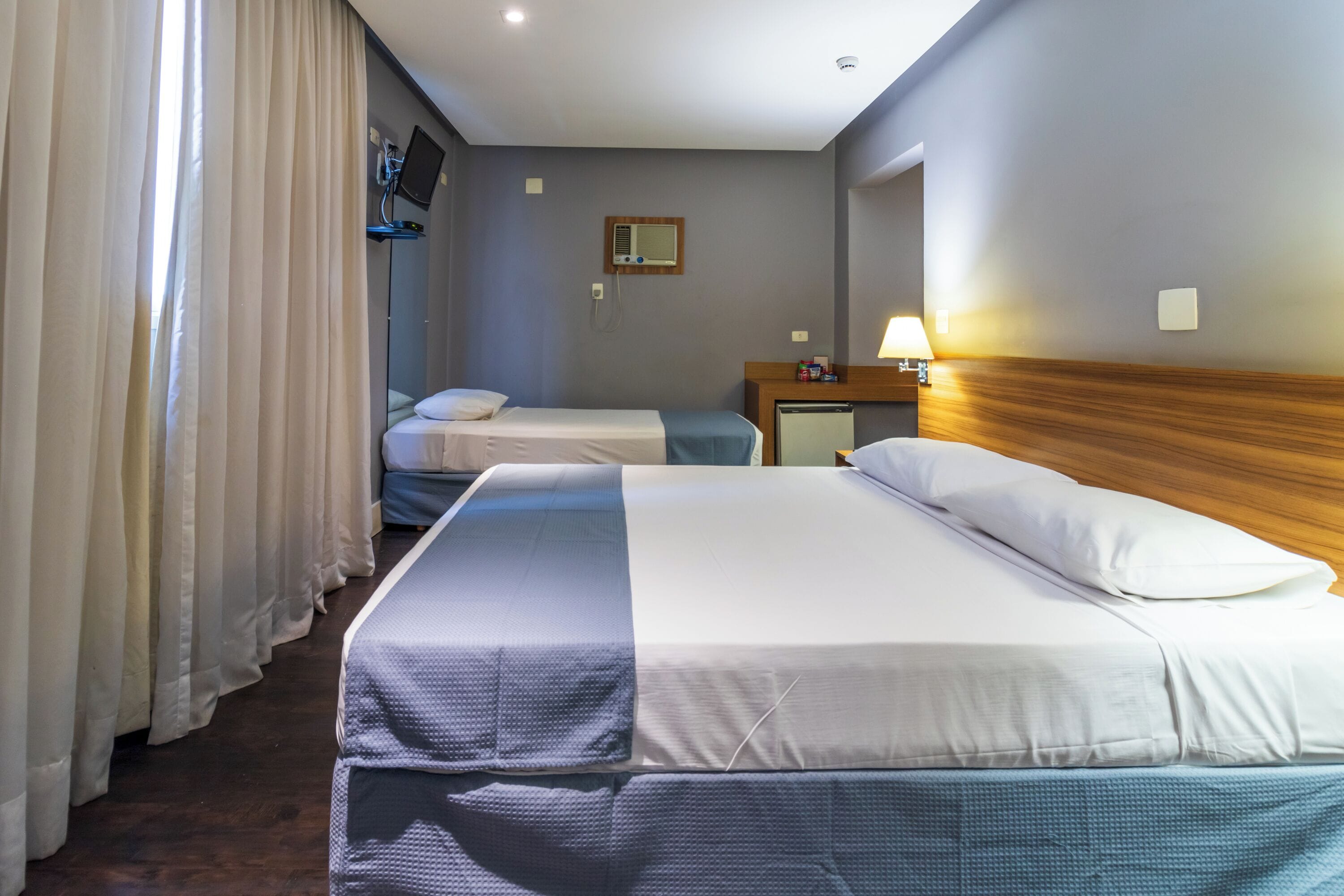A hotel room with two beds, a wooden headboard, a bedside table with a lamp, and a TV mounted on the wall.
