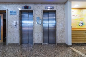 Due ascensori in un atrio con pavimento in marmo, un cartello con la scritta 'Masp Metropolitan' e un orologio digitale sopra l'ascensore a sinistra.