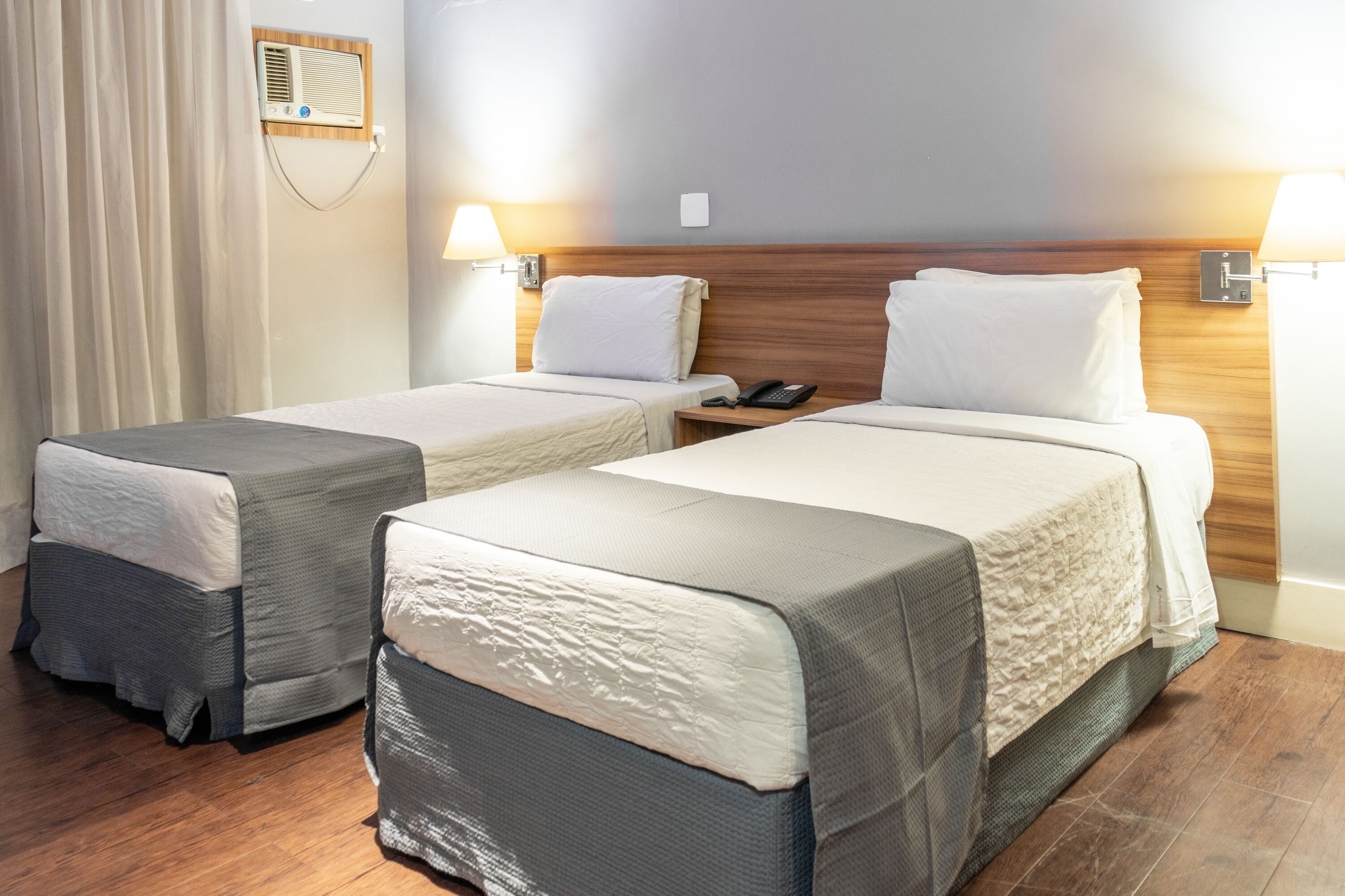 A hotel room with two beds, wooden headboards, bedside lamps, a telephone, and a wall-mounted air conditioner.