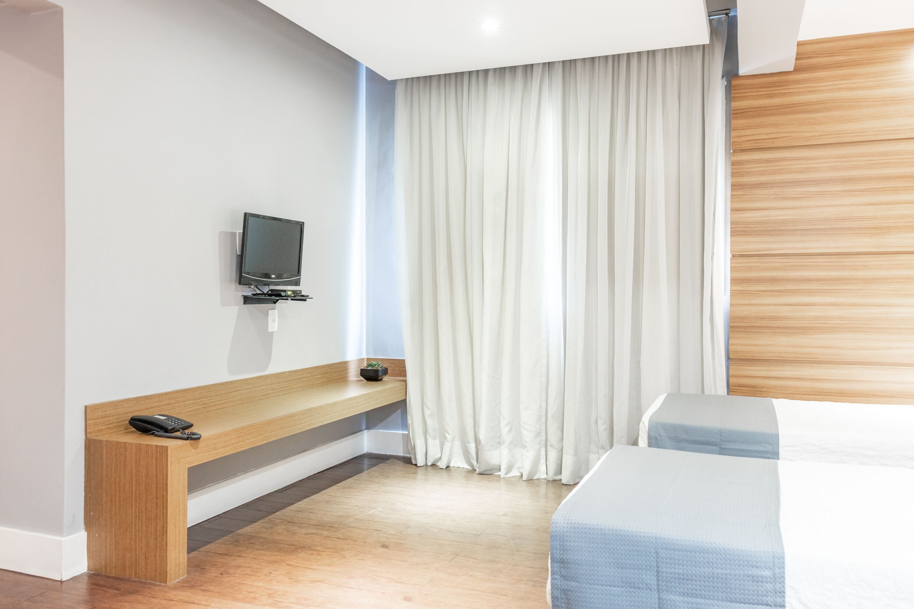 A hotel room with a wooden bench, a television, a bed, and a large window with curtains.