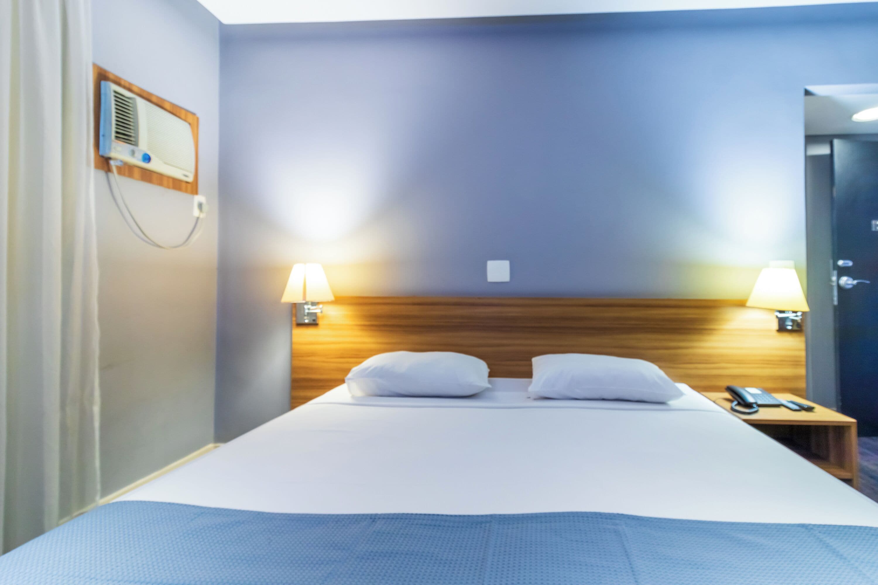 A hotel room with a bed, two bedside lamps, a wooden headboard, a nightstand with a phone, and a wall-mounted radio.
