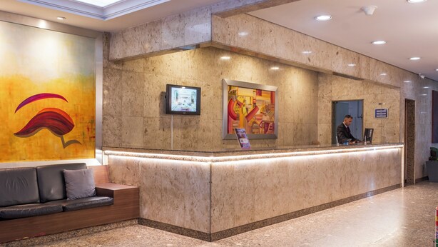 A hotel reception area with a marble counter, a sofa with cushions, a digital screen, and a colorful abstract painting on the wall. - Braston Augusta by Castelo Itaipava (São Paulo)