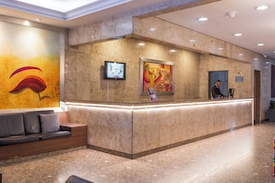Un'area reception di un hotel con un bancone in marmo, una poltrona con cuscini, uno schermo digitale e un quadro astratto dai colori vivaci appeso al muro.