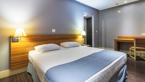 A hotel room with a large bed, two bedside lamps, a wooden headboard, a desk with a chair, and a door leading to another room. - Braston Augusta by Castelo Itaipava (São Paulo)