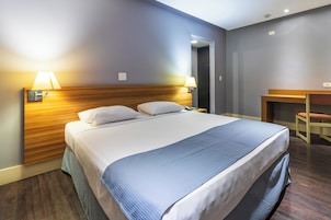 Una camera d'albergo con un letto grande, due lampade ai comodini, una testiera in legno, una scrivania con una sedia e una porta che dà su un'altra stanza.