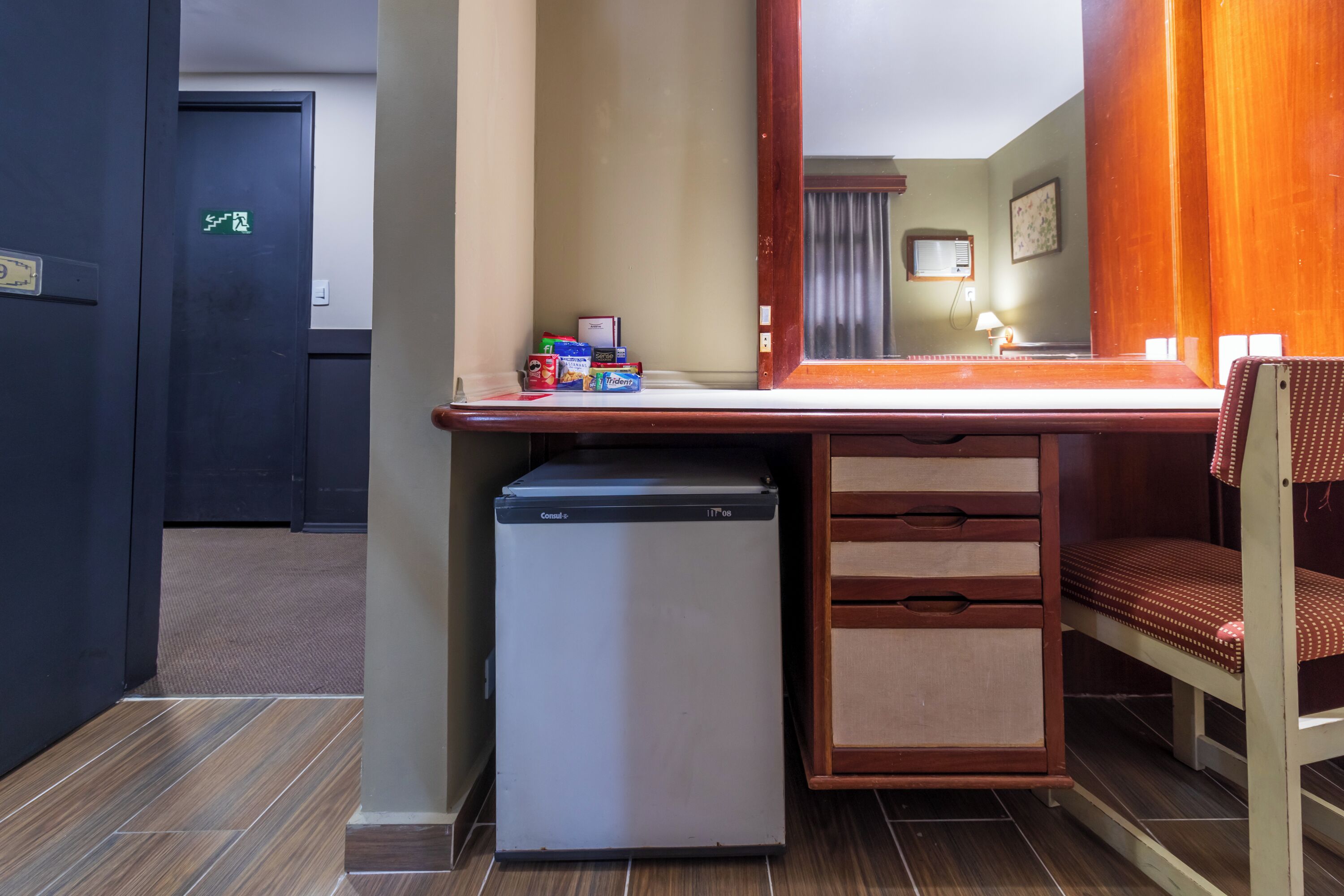 A compact hotel room with a kitchenette, a small refrigerator, a wooden desk, a chair, and a mirror.