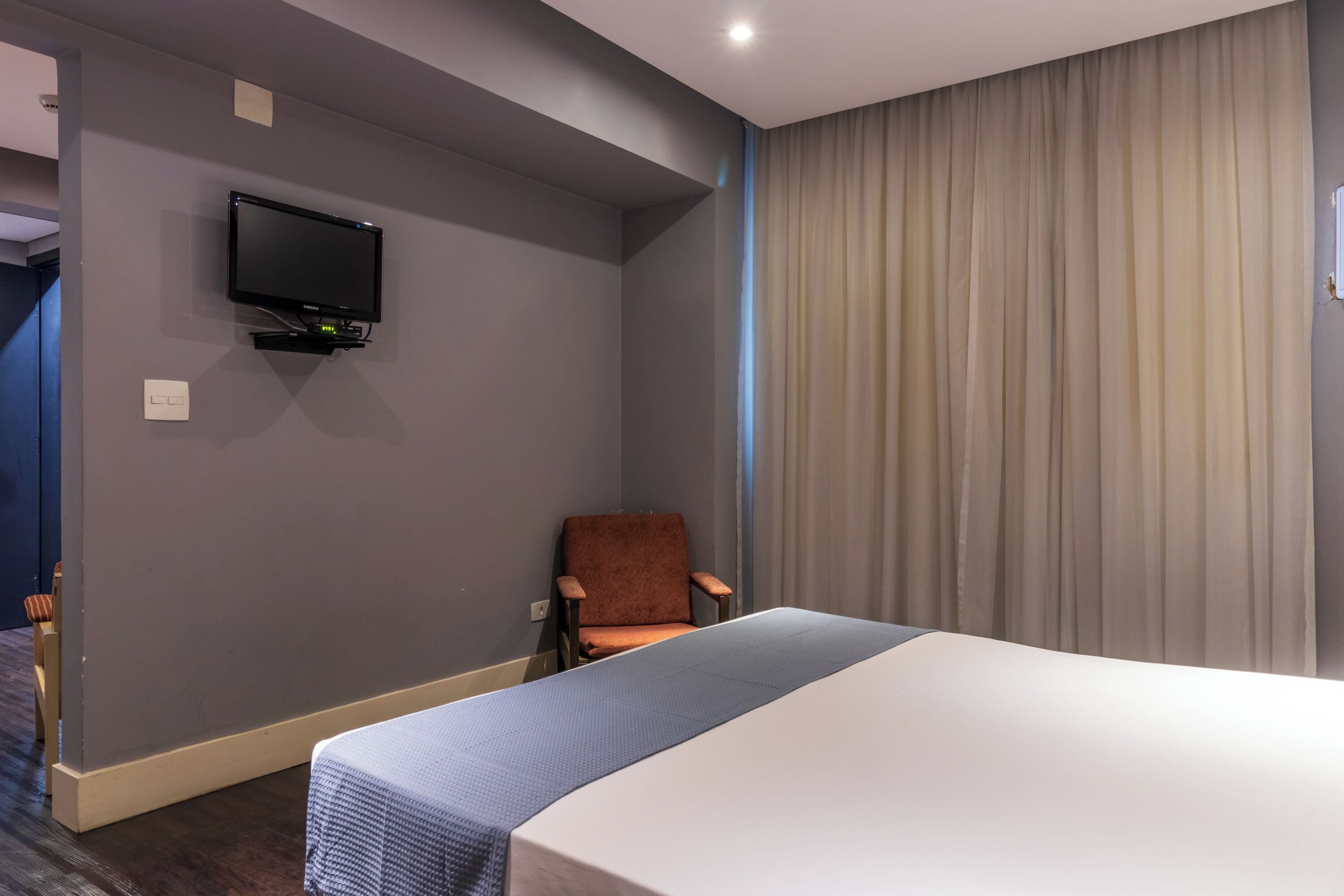 Una habitació d'hotel amb un llit, una cadira, un televisor fixat a la paret i una cortina.