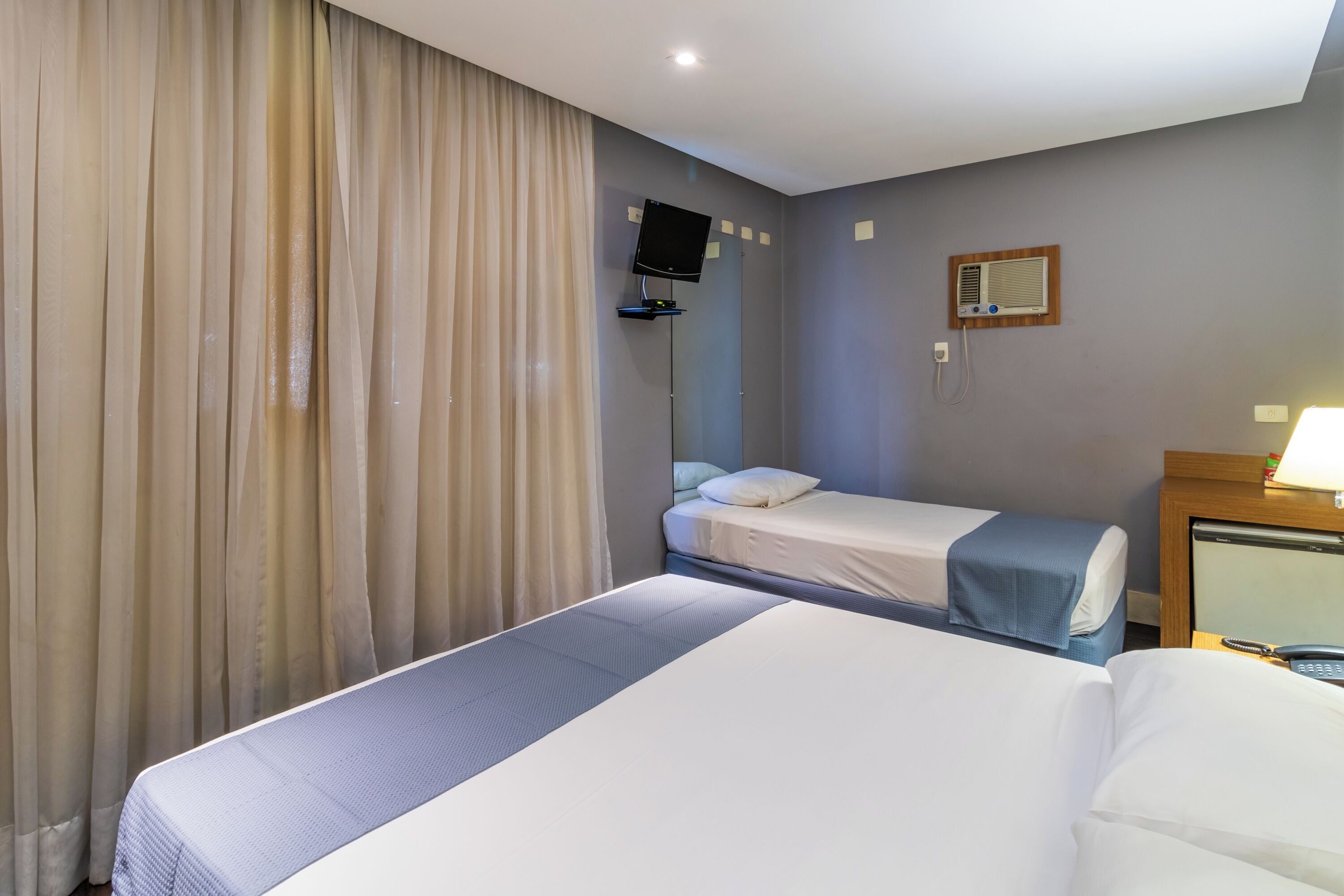 A hotel room with two beds, a TV mounted on the wall, an air conditioning unit, and a bedside table with a lamp.