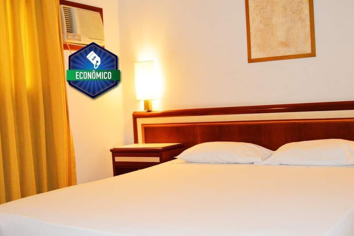Quarto de hotel com cama, mesa de cabeceira, luminária, ar condicionado fixado na parede e um quadro emoldurado.