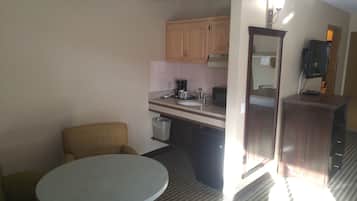 Habitación de hotel pequeña con una mesa redonda, una sola silla, kitchenette con cafetera y un espejo.