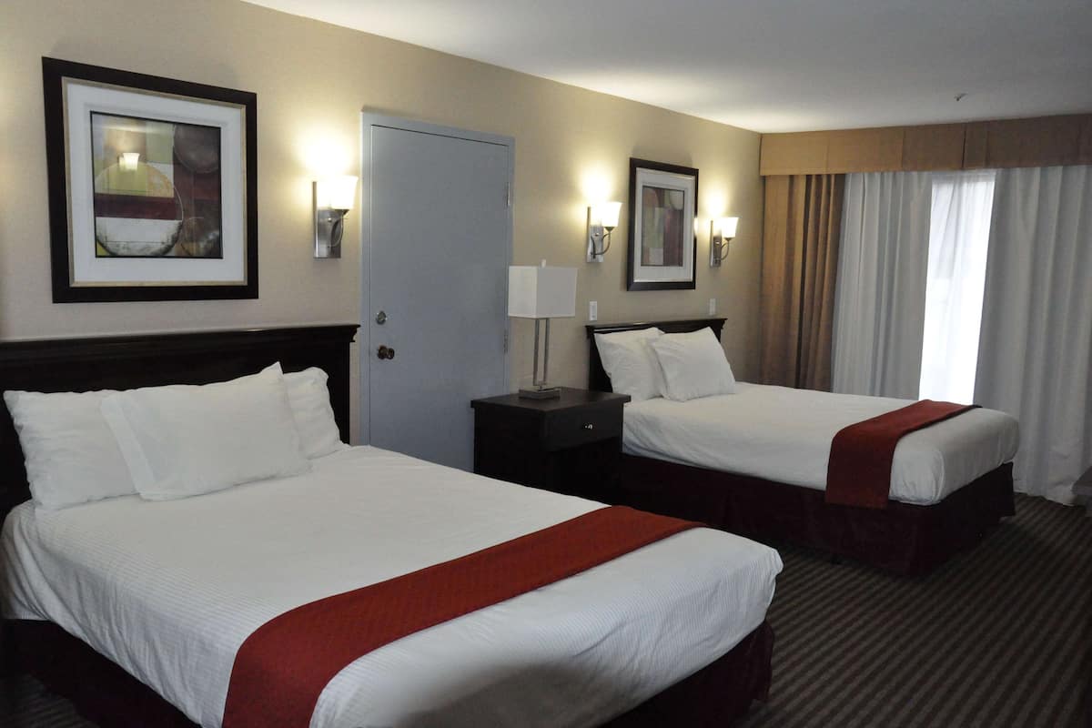 A hotel room with two beds, a door, a nightstand with a lamp, and framed artwork on the walls.