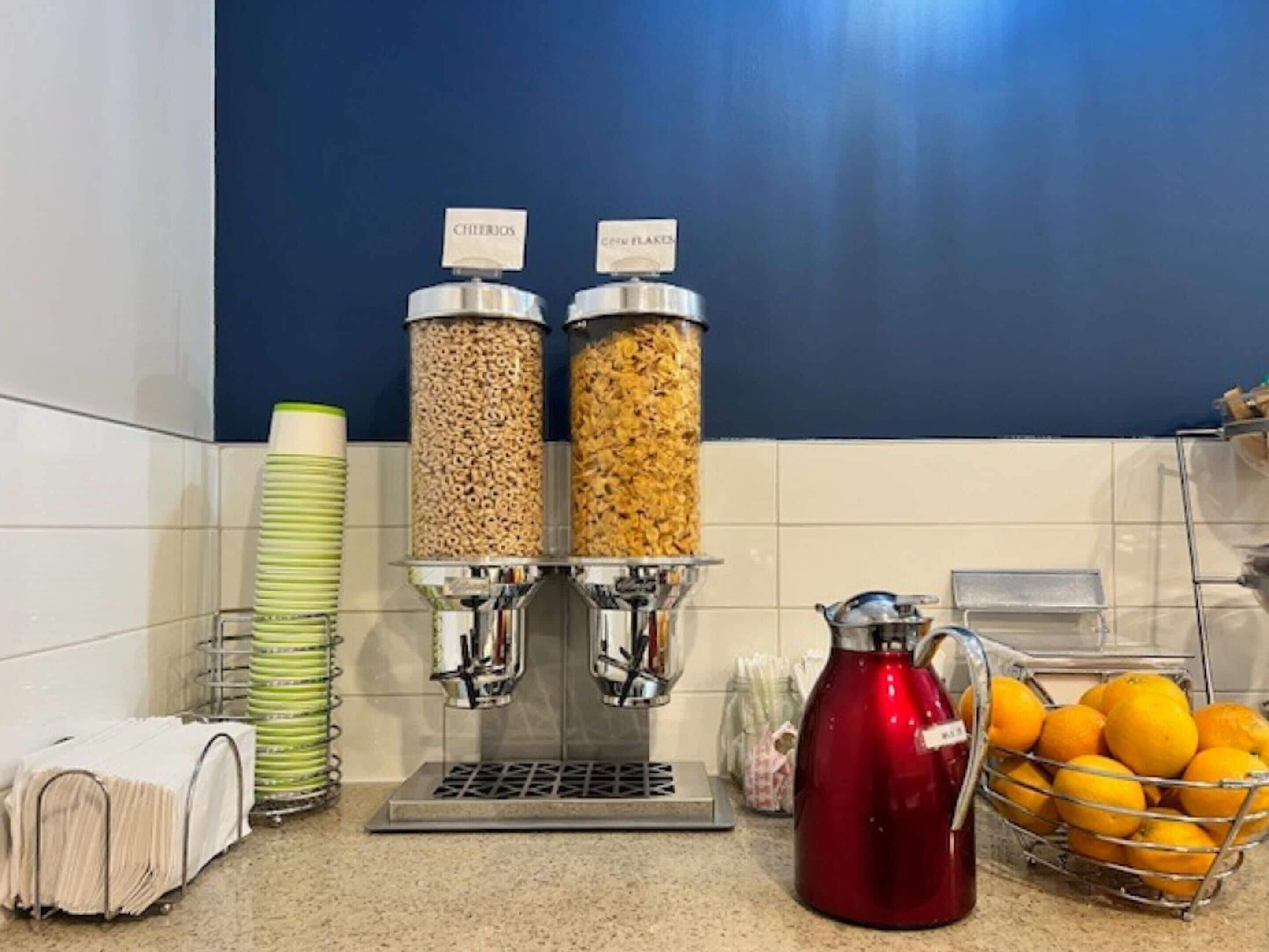 Una zona de cocina con cafetera, dispensadores de cereales, hervidor de agua y una cesta de naranjas.