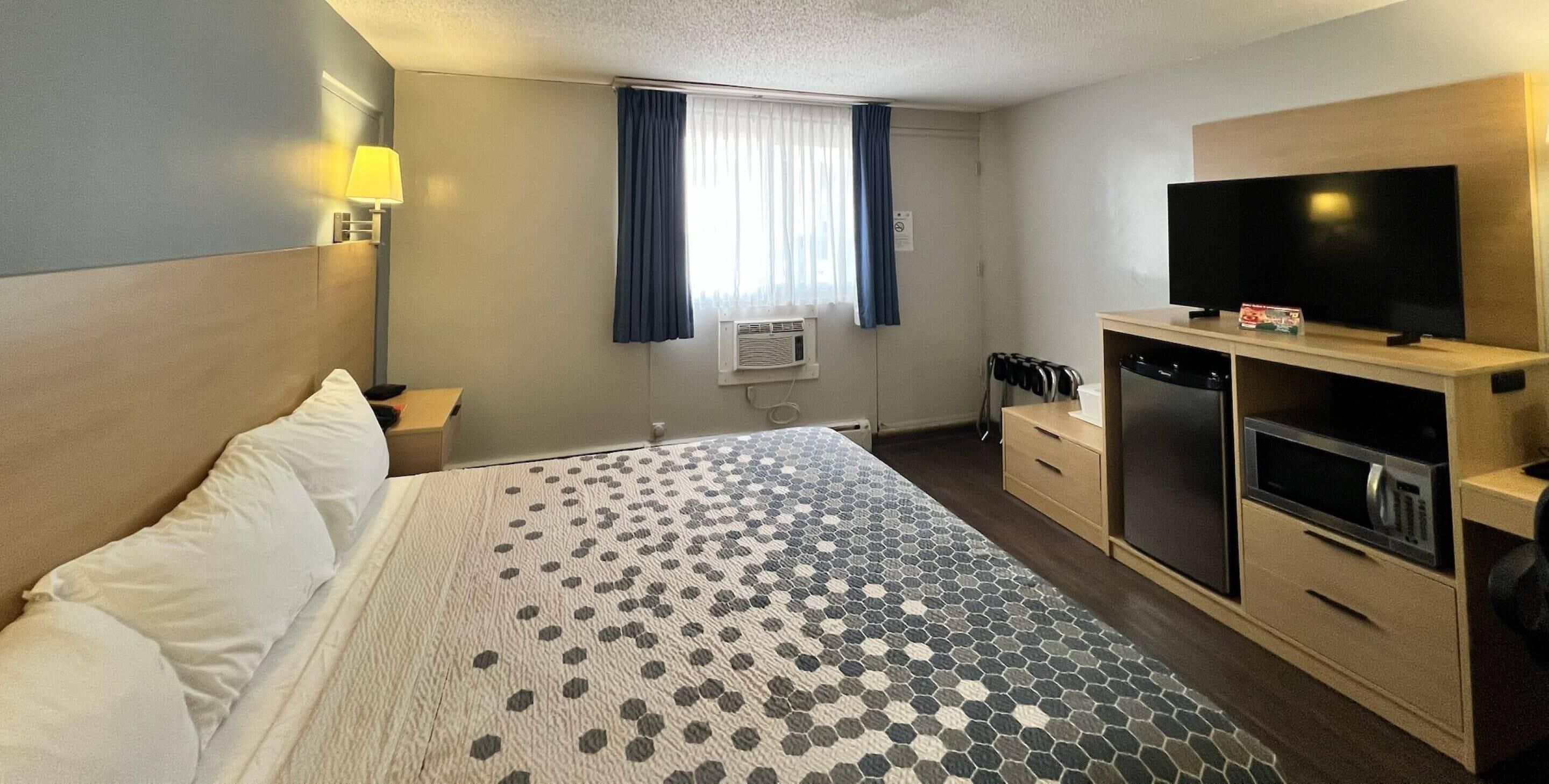 Habitación de hotel con cama, televisor, refrigerador pequeño, microondas y ventana con cortinas.
