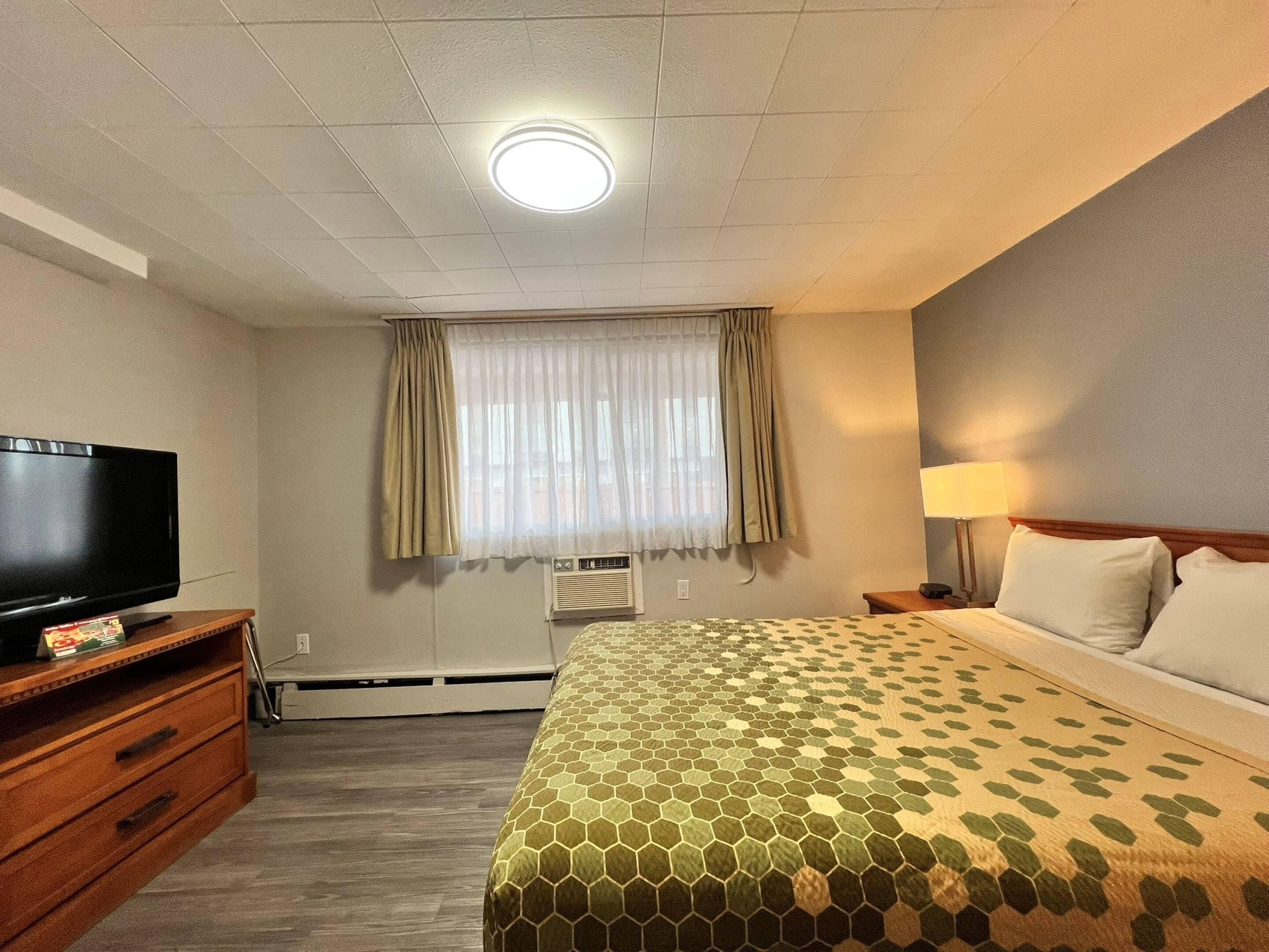 Habitación de hotel con televisión de pantalla plana, cama con colcha estampada, lámpara de noche y ventana con cortinas.