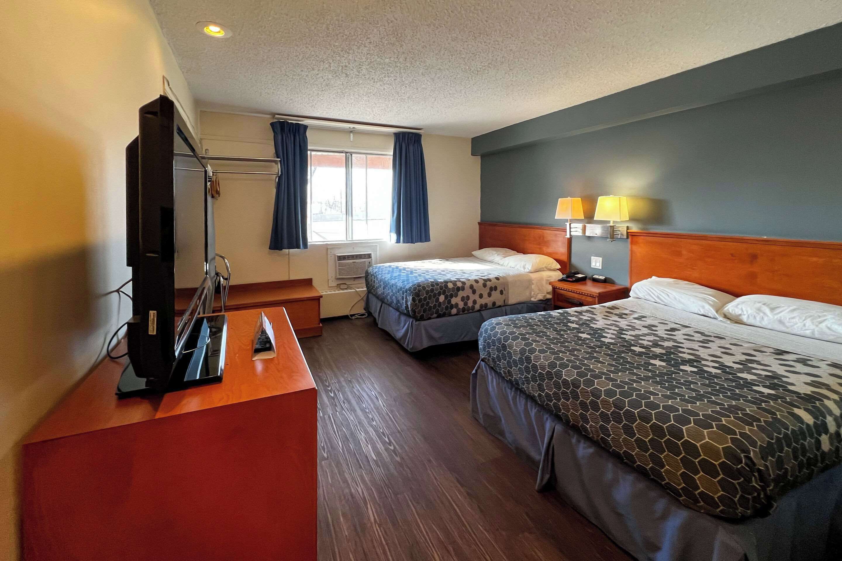 A hotel room with two beds, a desk with a television, a window with curtains, and a lamp on the nightstand.