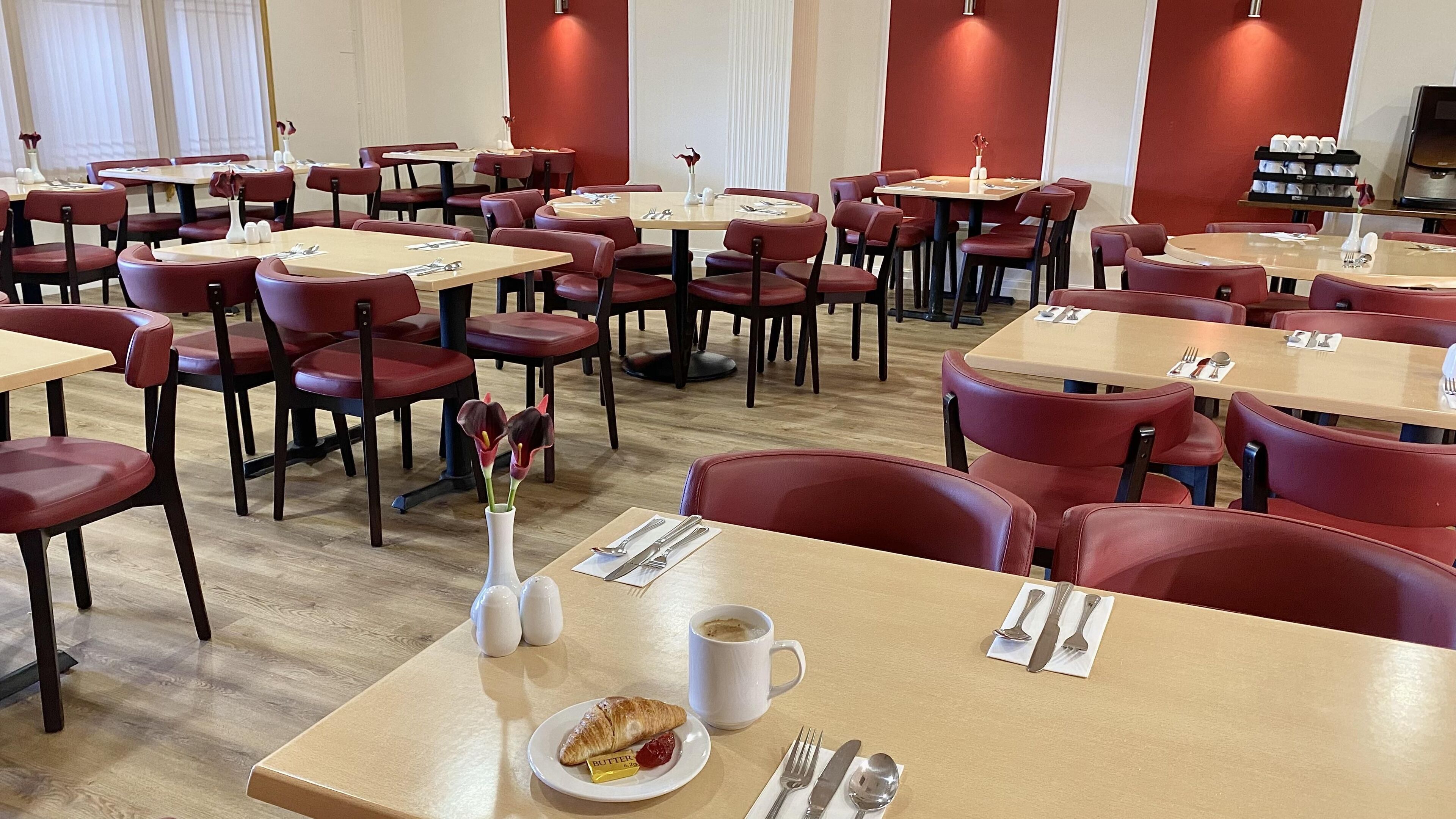 A hotel dining area with multiple tables and chairs, each set with cutlery and a small vase with flowers.