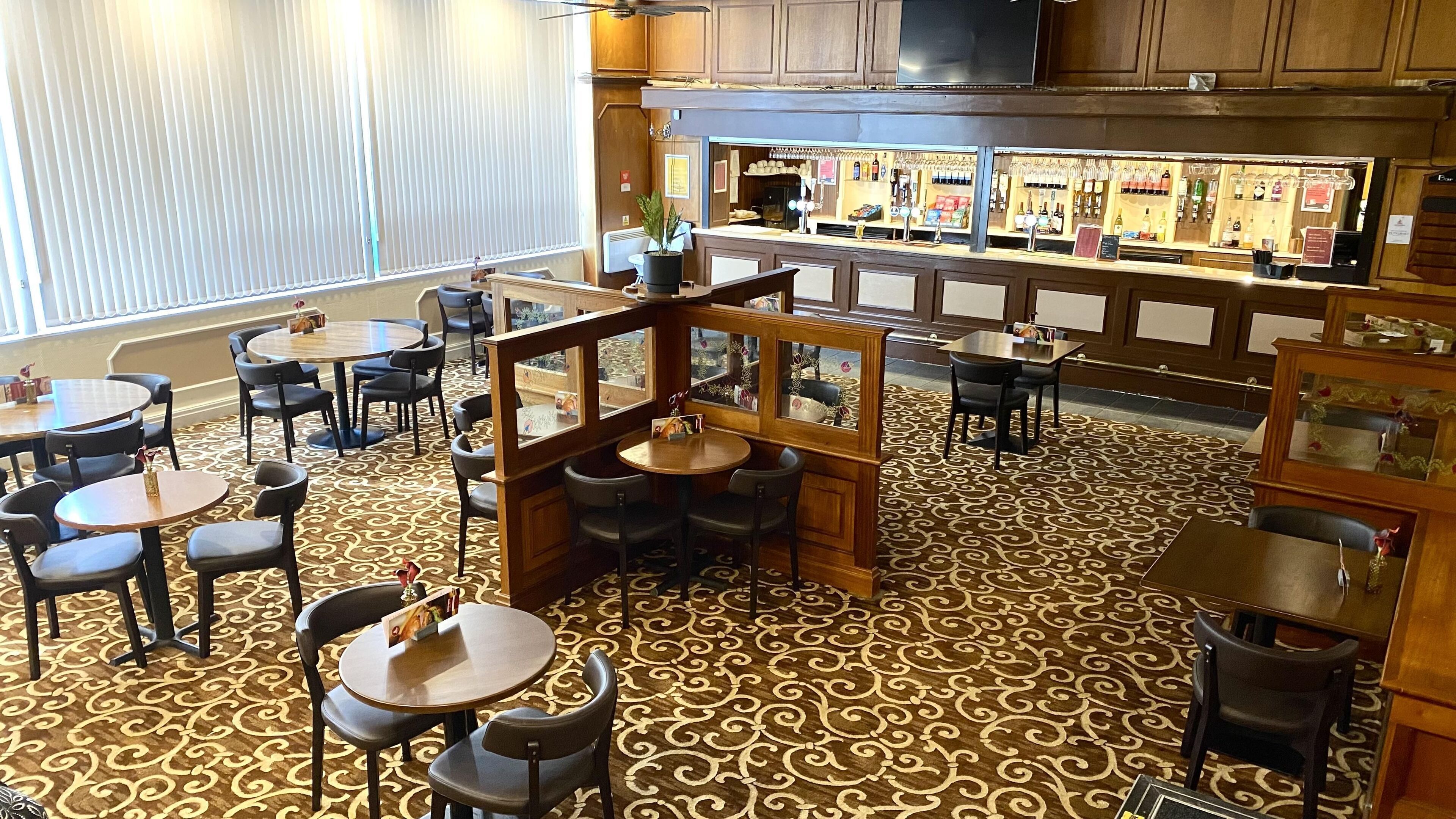 A restaurant with round tables and chairs, a bar area with shelves of bottles, and a patterned carpet.