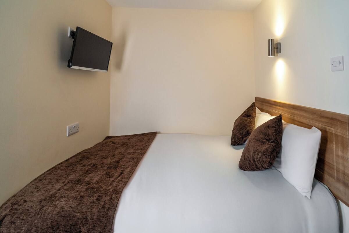 A neatly made bed with a wooden headboard, a mounted flat-screen TV, and a wall-mounted light fixture.