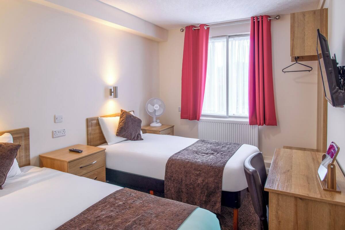 A hotel room with two beds, a bedside table with a lamp, a small table with a mirror, a TV mounted on the wall, and a window with red curtains.