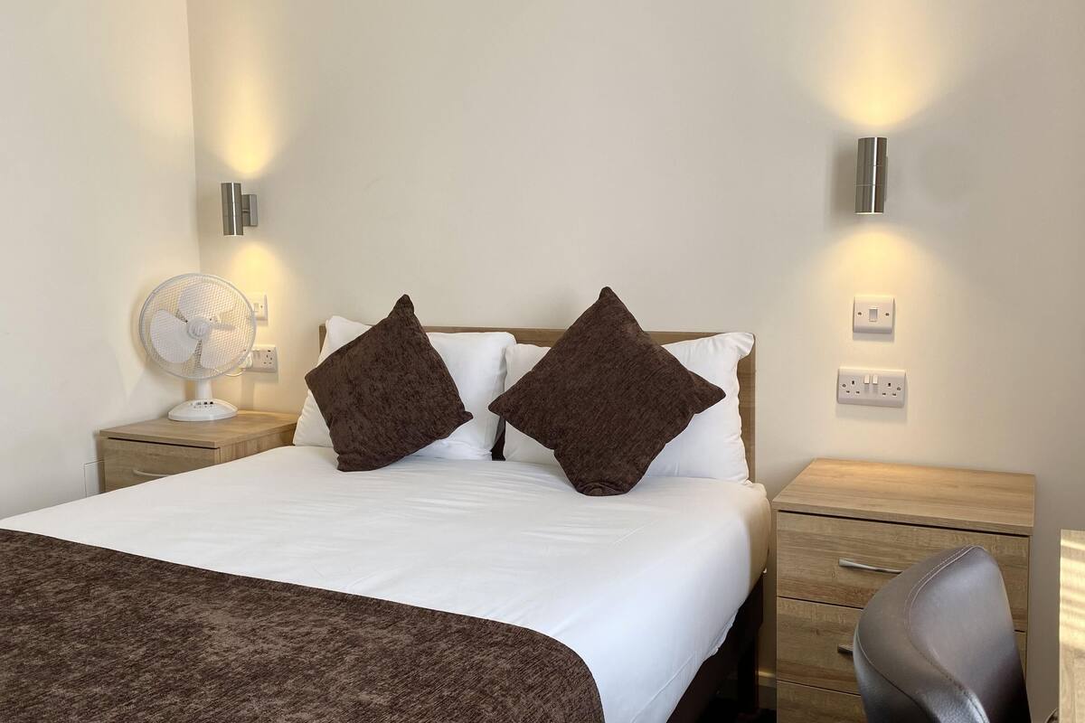 A hotel room with a bed, two bedside tables, a standing fan, and a desk with a chair.