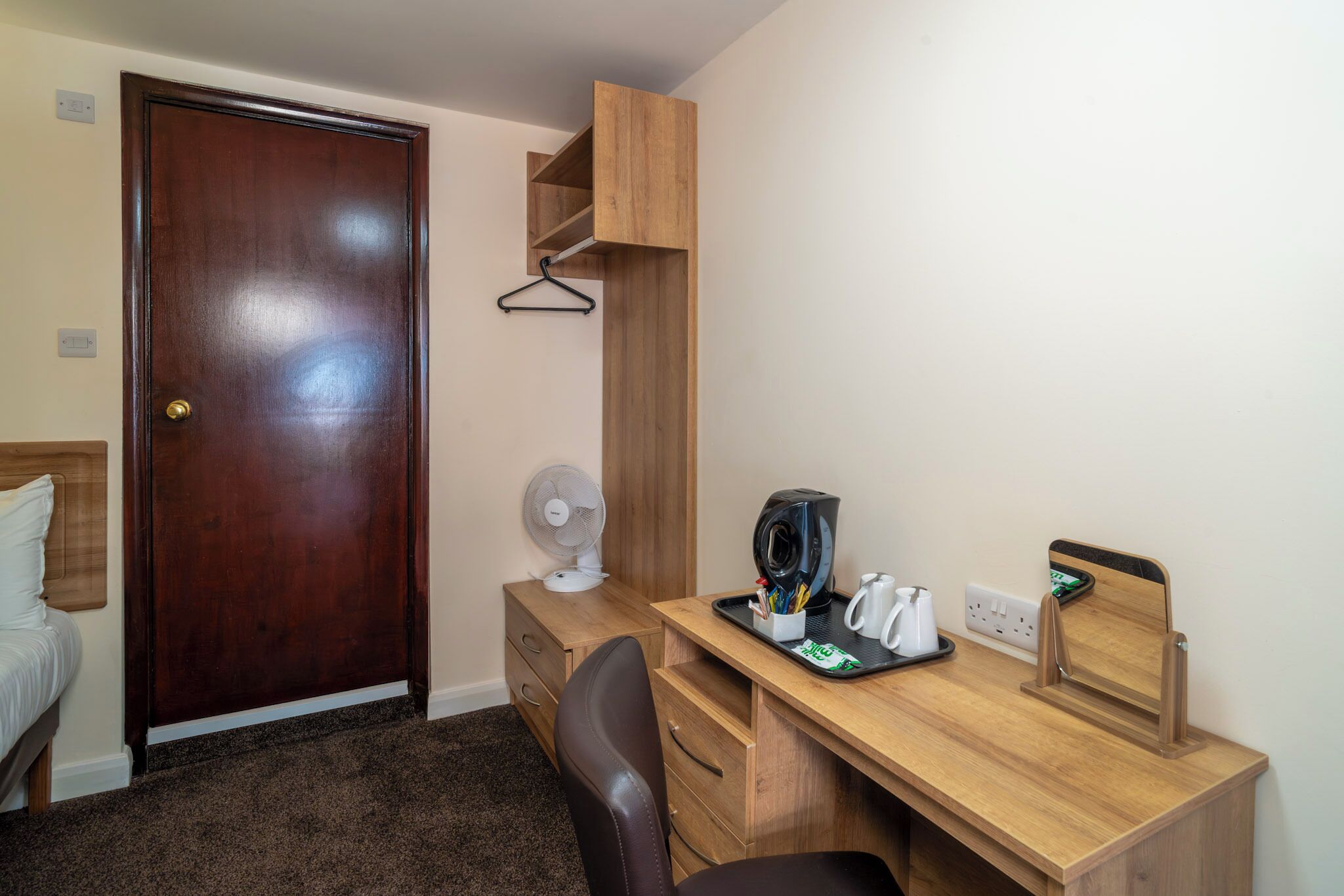 Habitación de hotel pequeña con un escritorio de madera, una silla, una cama, un armario y un pequeño ventilador.