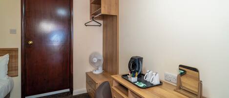 Habitación de hotel pequeña con un escritorio de madera, una silla, una cama, un armario y un pequeño ventilador.