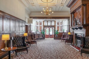 A grand lobby with a patterned carpet, leather armchairs, a chandelier, and a fireplace. - Russ Hill Hotel (Horley)