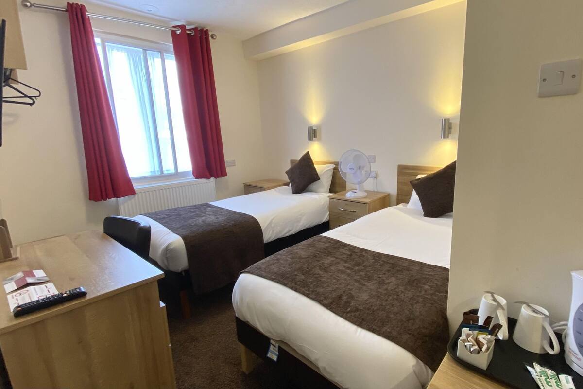 A hotel room with two beds, a desk, a chair, a TV, a window with curtains, and a bedside table with a lamp and a fan.