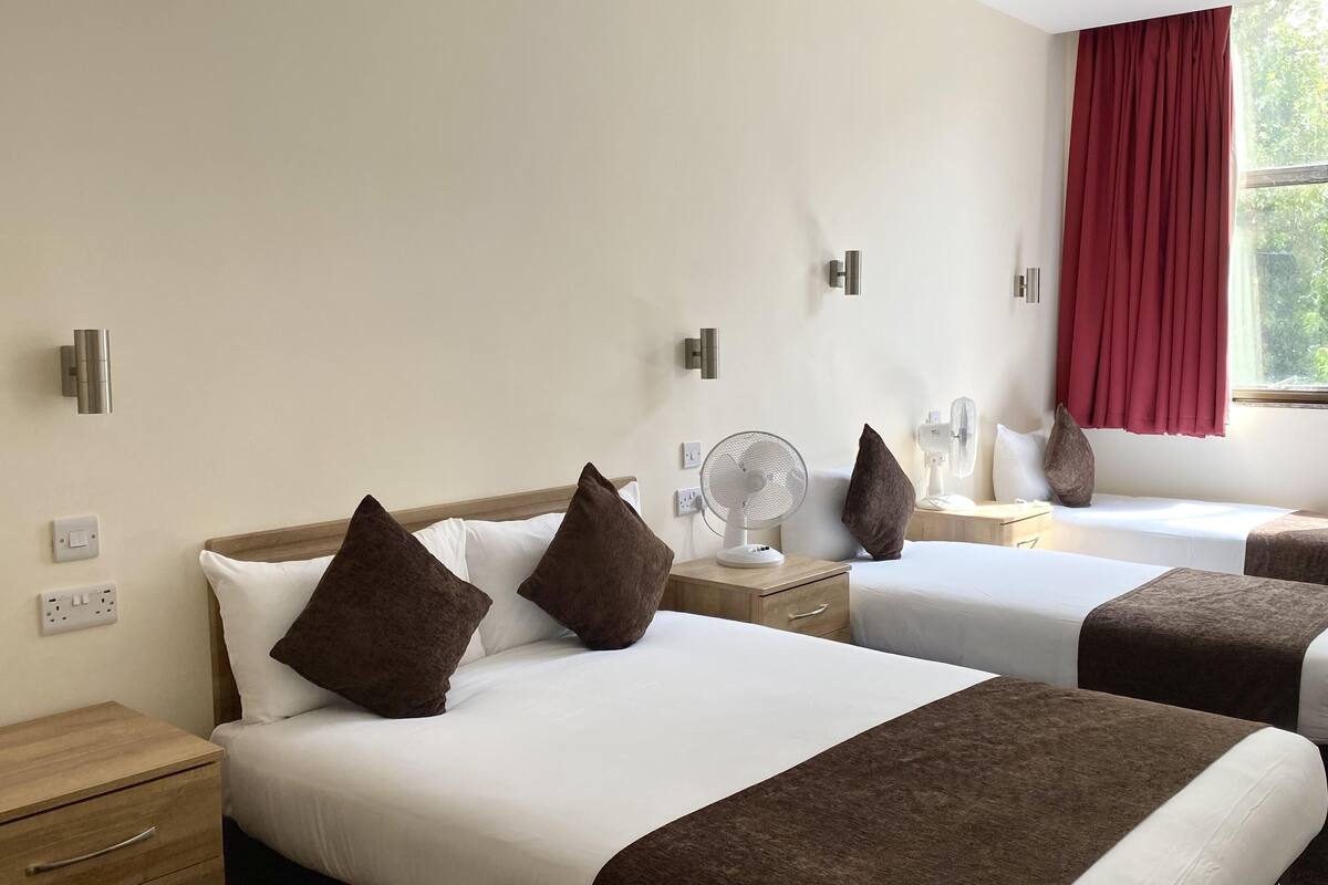 A hotel room with two beds, each with a white mattress and brown headboard, a bedside table with a fan, and a window with red curtains.