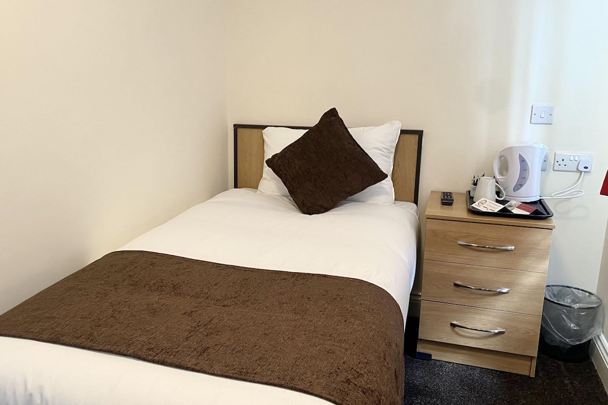 A hotel room with a bed, a bedside table with a kettle, a trash bin, and a drawer unit.