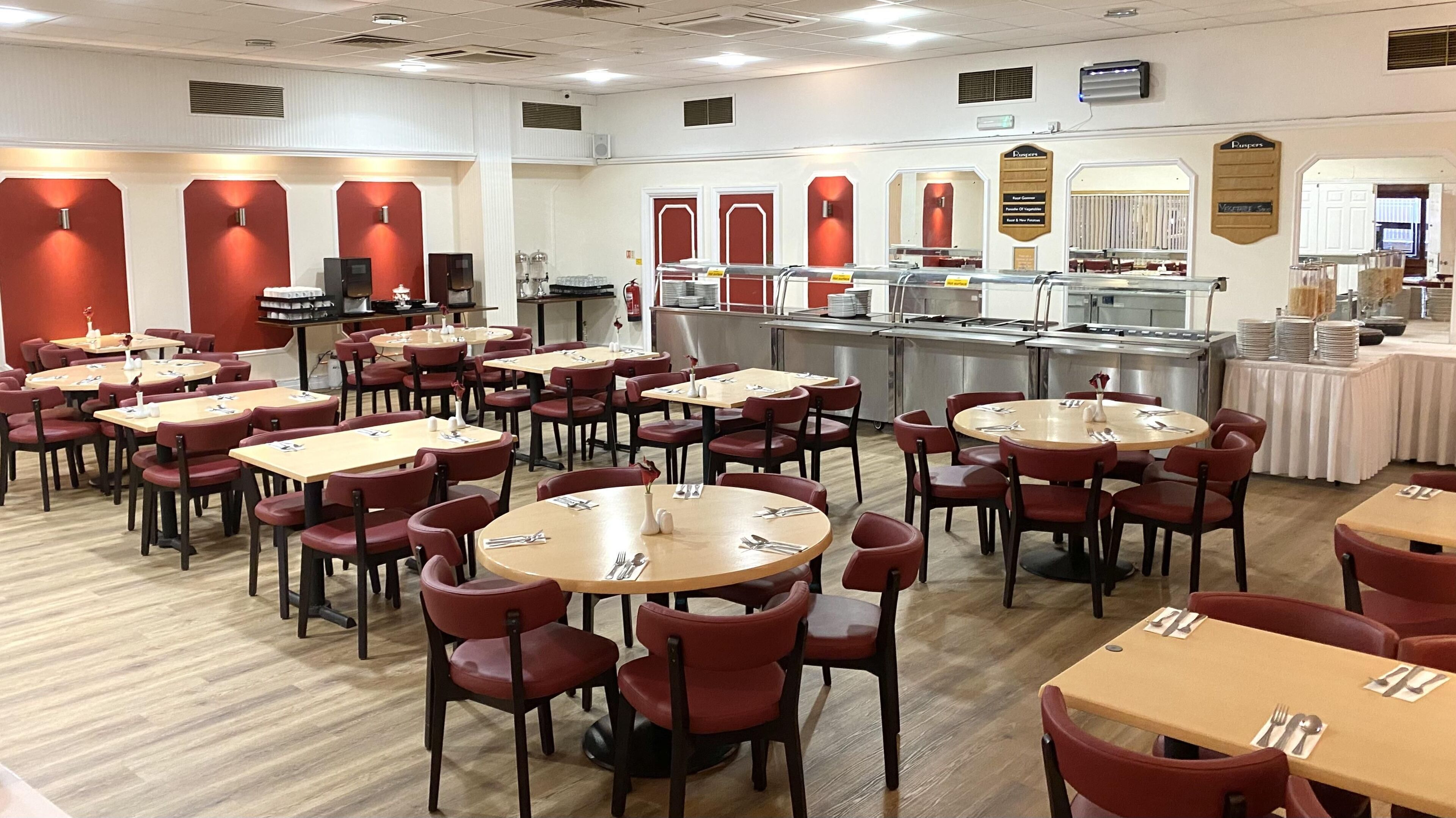 A dining area with multiple tables and chairs, a buffet line, and a kitchen area in the background.