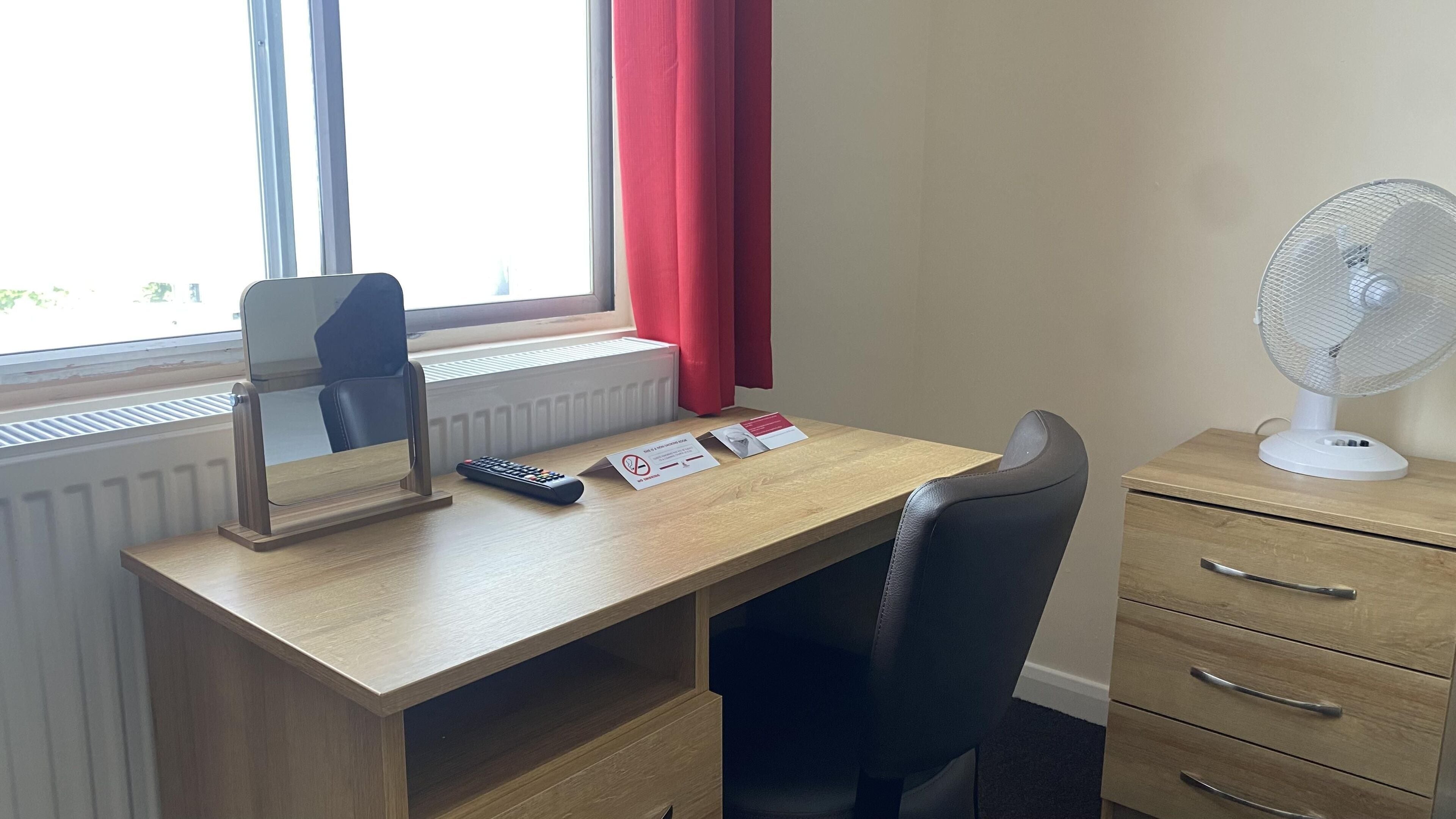 A hotel room with a wooden desk, a chair, a white standing fan, a mirror, a remote control, and a set of cards on the desk.