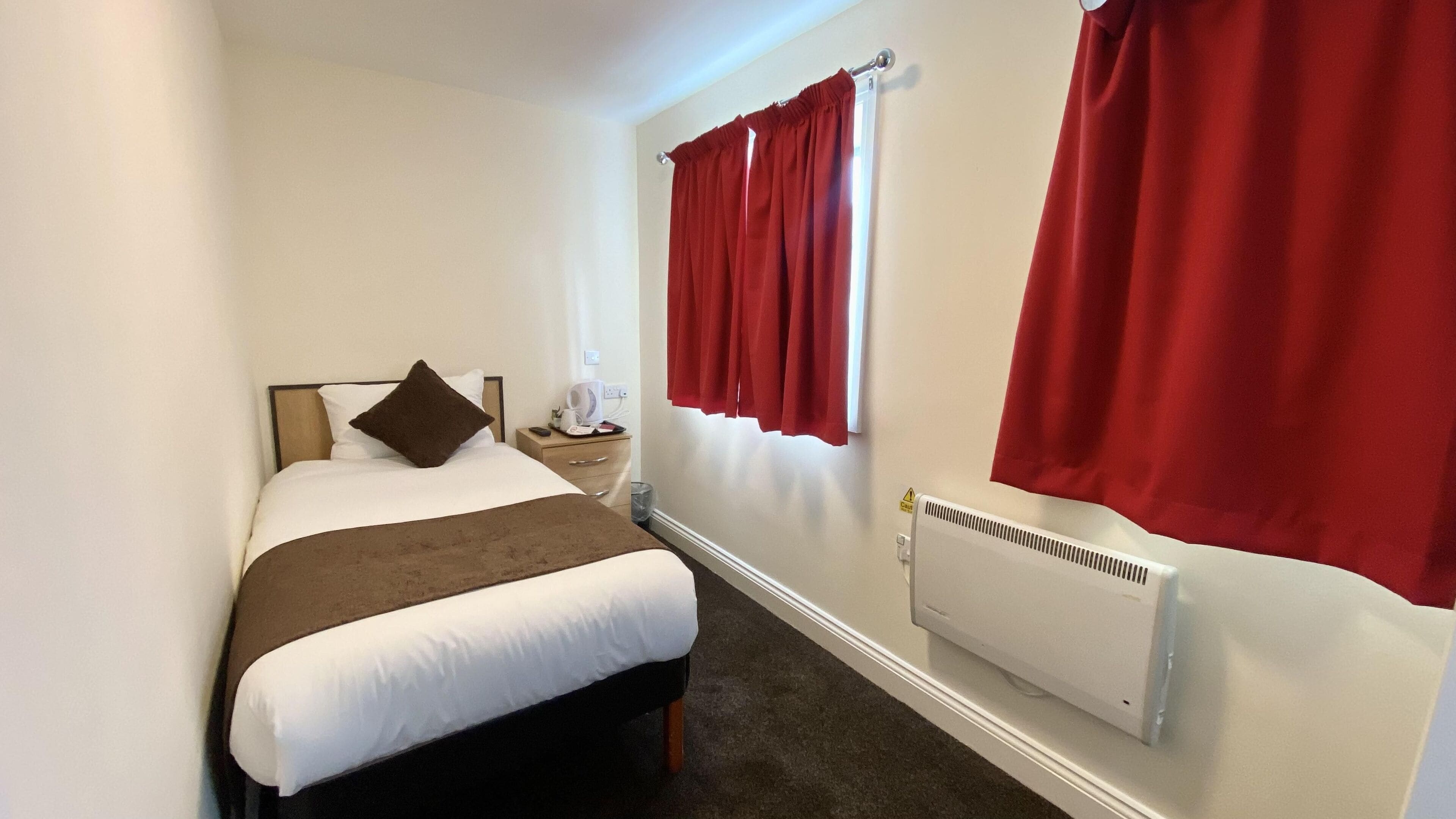 A hotel room with a bed, a nightstand, a window with red curtains, and a radiator.