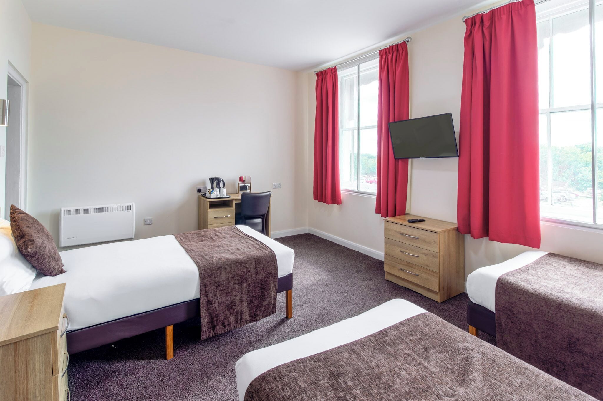 A hotel room with two beds, a desk, a chair, a TV, and a window with red curtains.