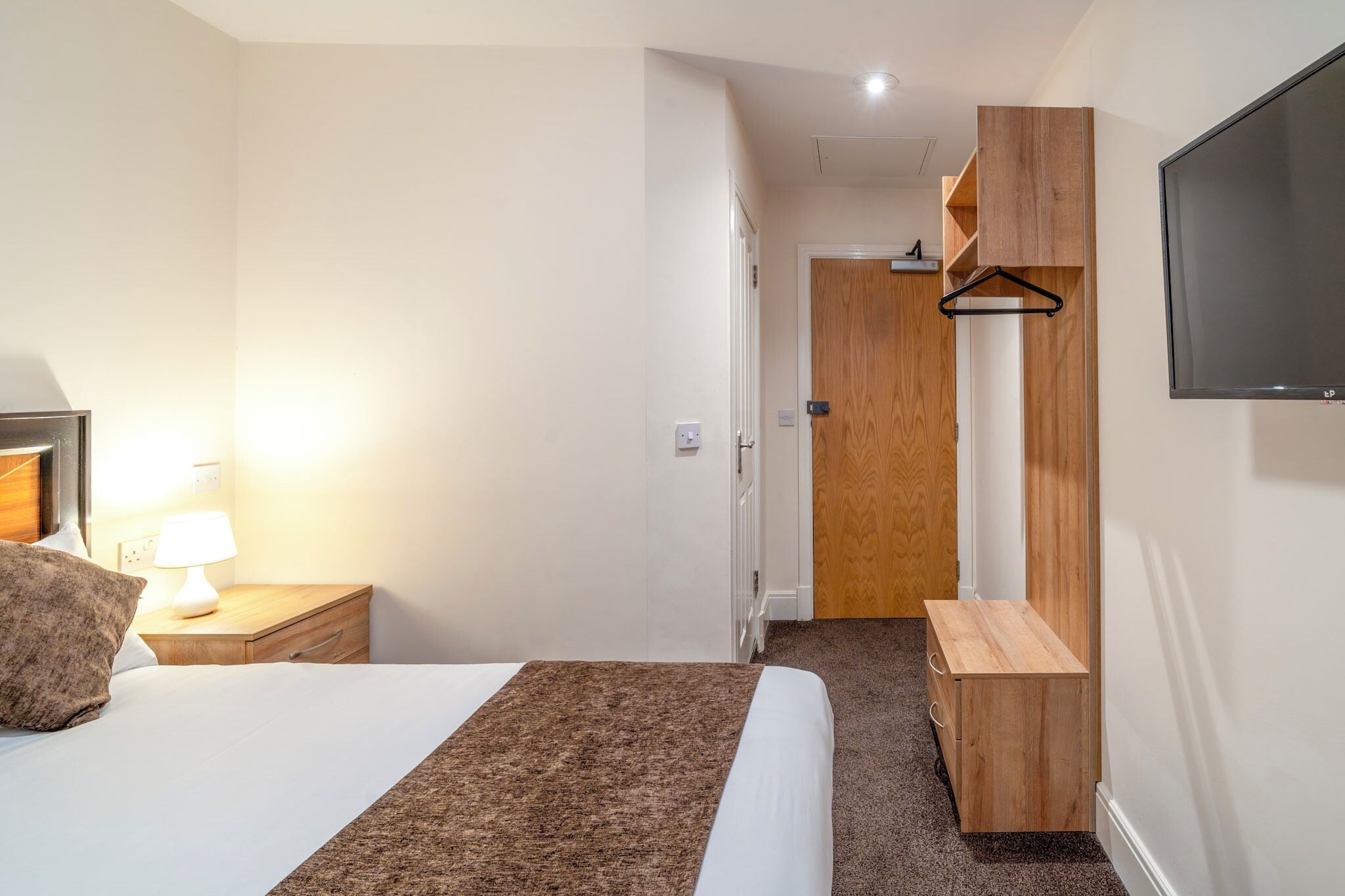 A hotel room with a bed, bedside table with a lamp, a wooden dresser, and a TV mounted on the wall.
