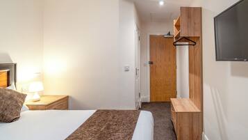 A hotel room with a bed, bedside table with a lamp, a wooden dresser, and a TV mounted on the wall.