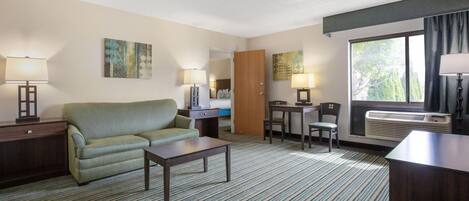 Une chambre d’hôtel avec un canapé, une petite table, un bureau, une chaise, une lampe et une fenêtre avec des rideaux.