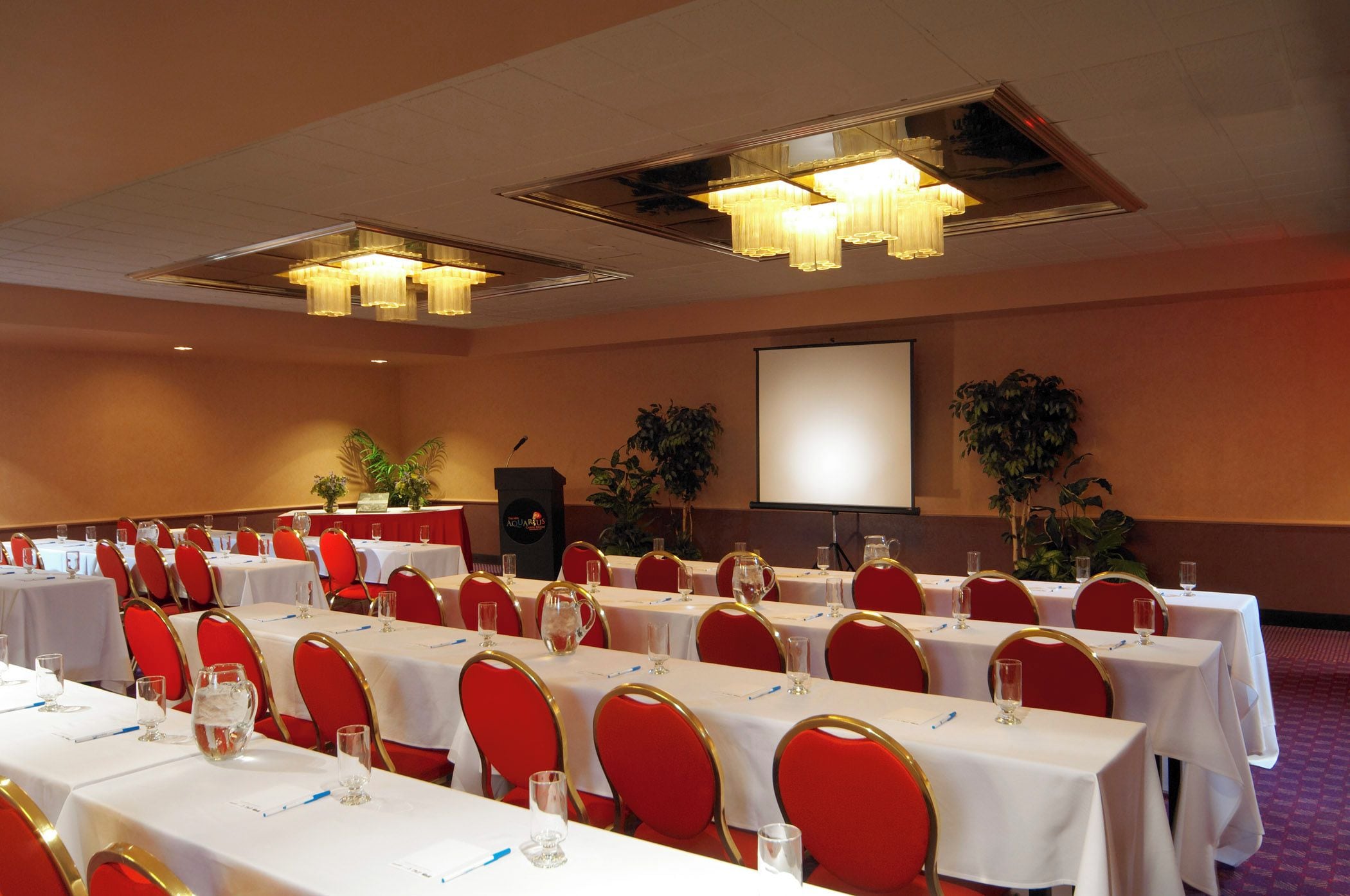 A conference room with rows of tables, red chairs, and a projector screen.