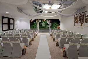 A wedding venue with rows of chairs, a white aisle, and a backdrop with greenery and floral arrangements.