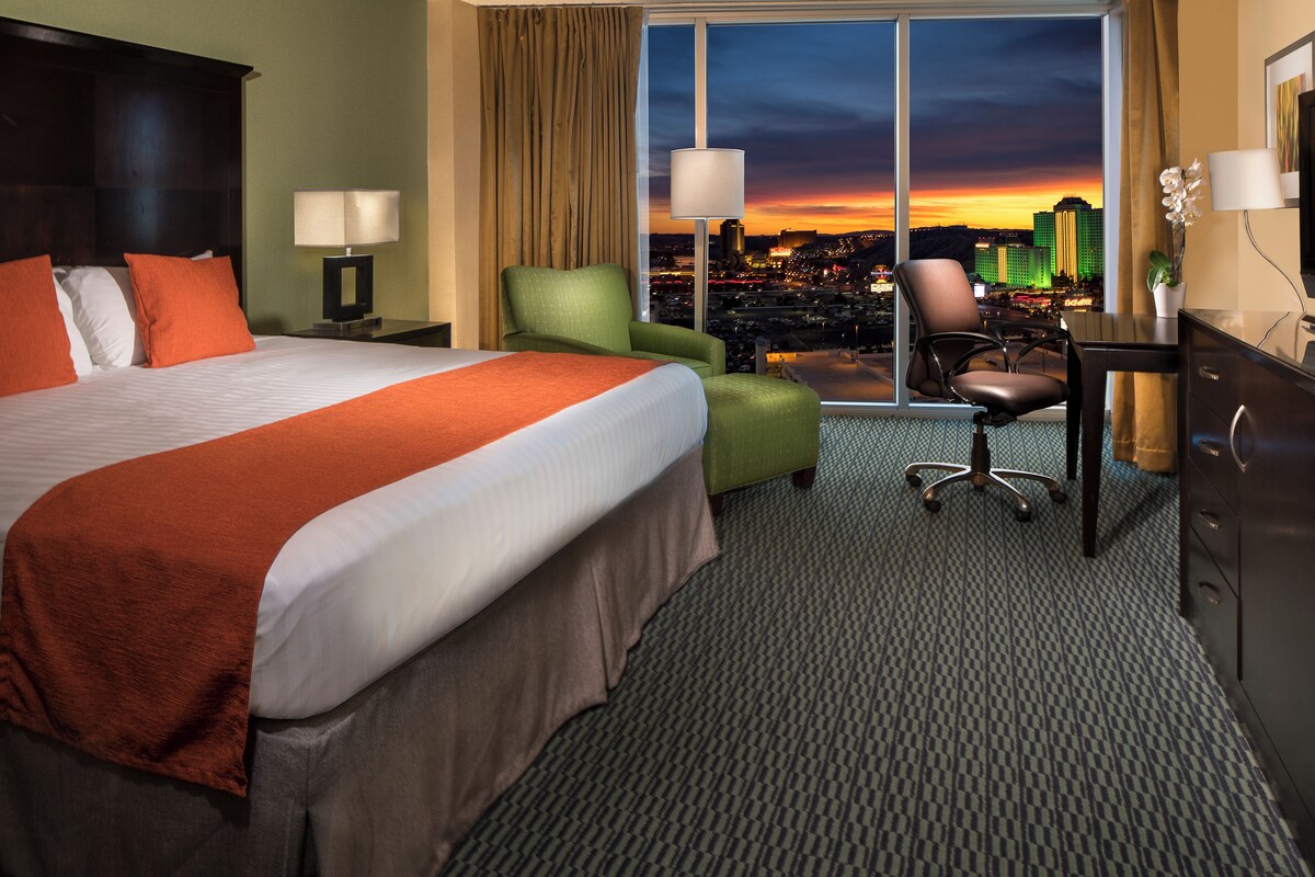 A hotel room with a large bed, a desk, a green armchair, and a view of a cityscape at sunset.