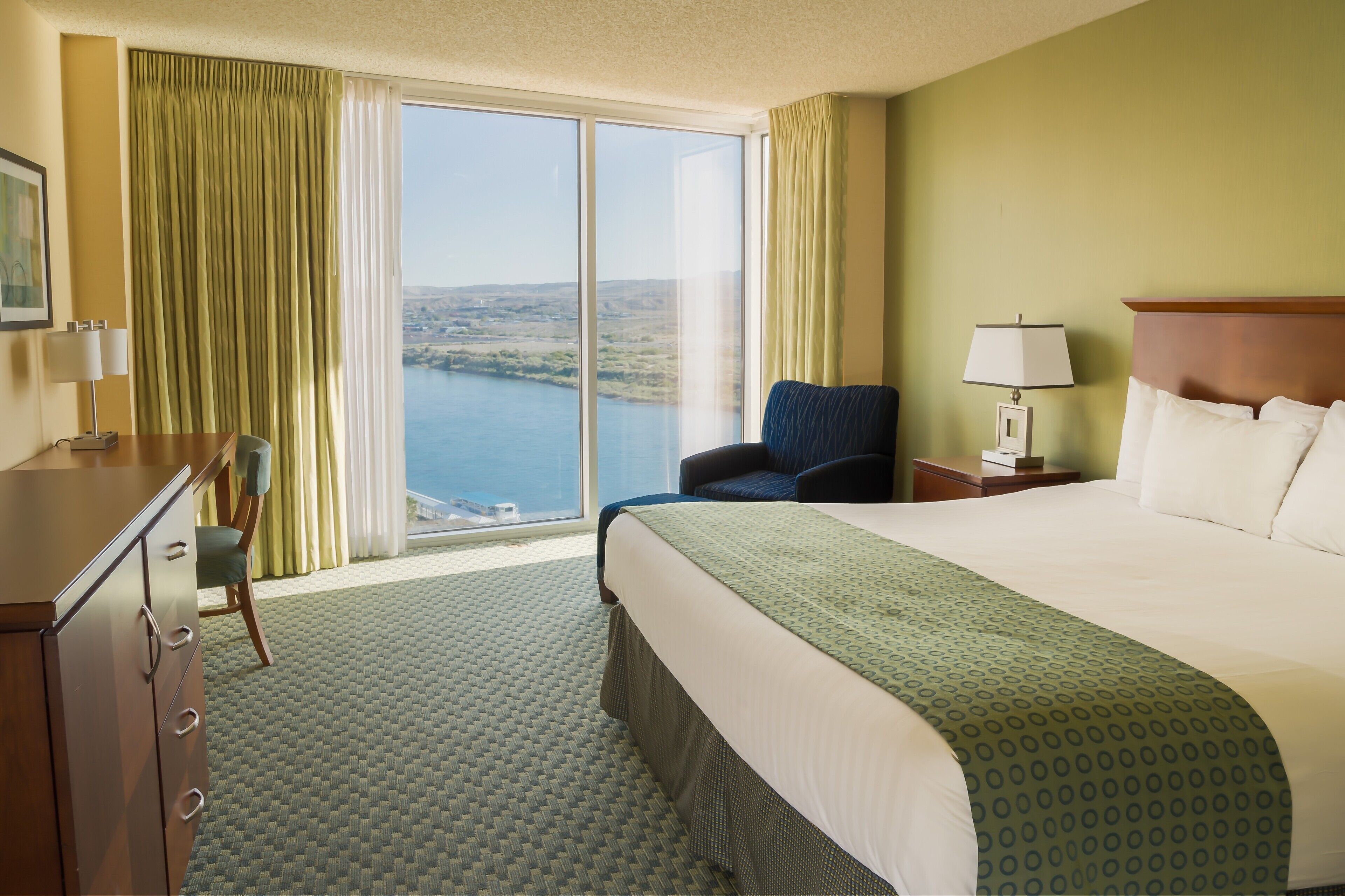 Une chambre d’hôtel avec un grand lit, un bureau, une chaise, une table de chevet et une vue sur une étendue d’eau, visible à travers une grande fenêtre.